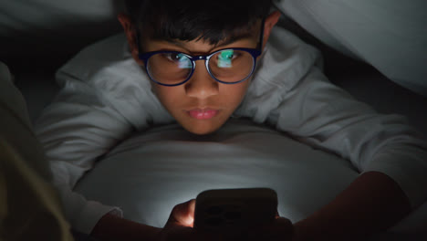 Close-Up-Of-Young-Boy-In-Bedroom-At-Home-Using-Mobile-Phone-To-Text-Message-Under-Covers-Or-Duvet-At-Night